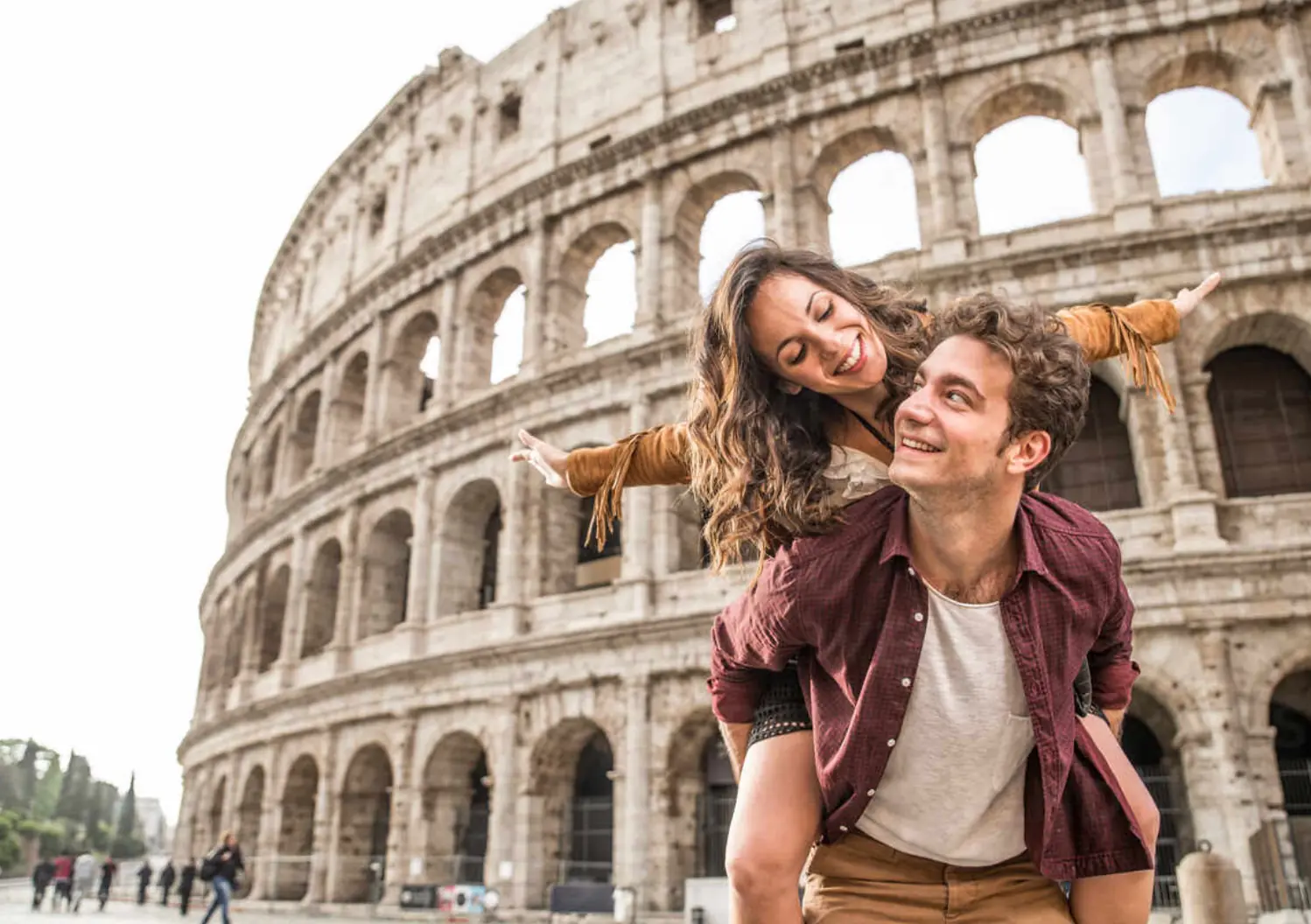 A laughing couple play in front of the arches of the Colosseum in Rome, the woman riding on the man's back with her arm outstretched.