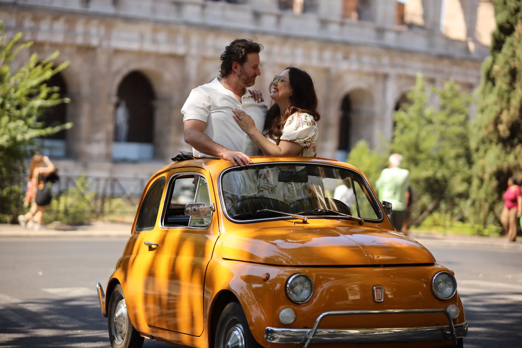 A couple stands up through the open roof of a bright orange vintage Fiat 500, laughing face to face, with the arches of the Colosseum blurred behind them.