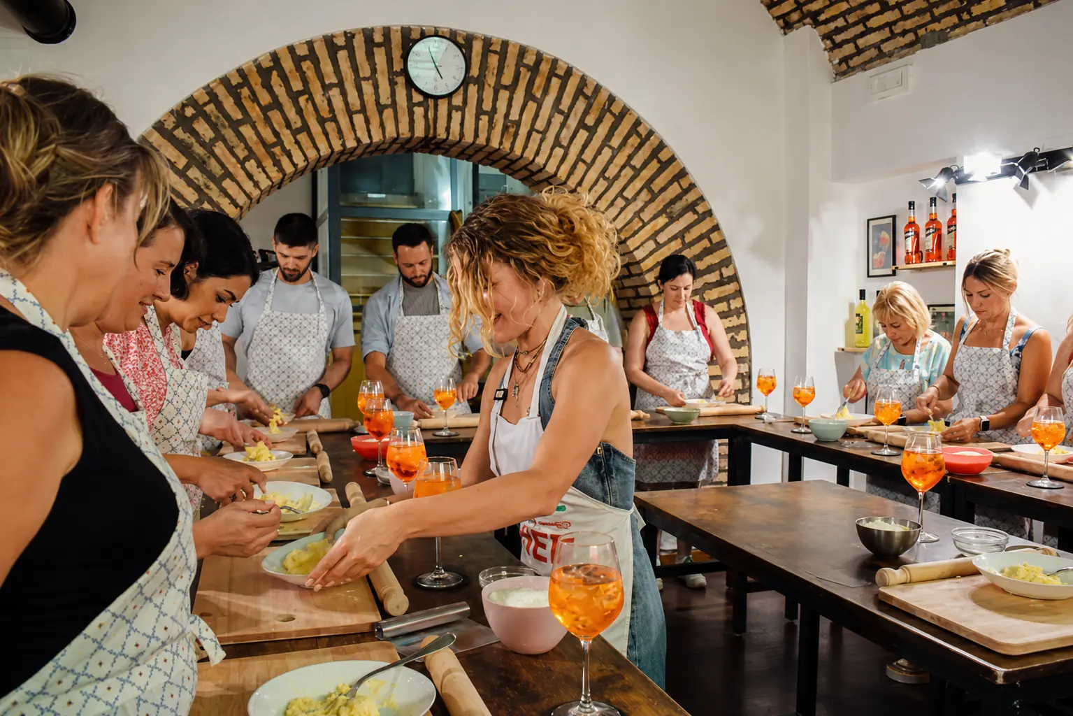 An instructor in an apron shapes pasta dough at a long wooden table while guests in matching aprons roll and knead alongside her under a rustic brick arch.