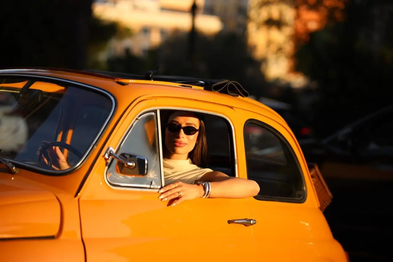 A woman in sunglasses leans out of the window of an orange vintage Fiat 500, lit by low golden sunlight on a Roman street.