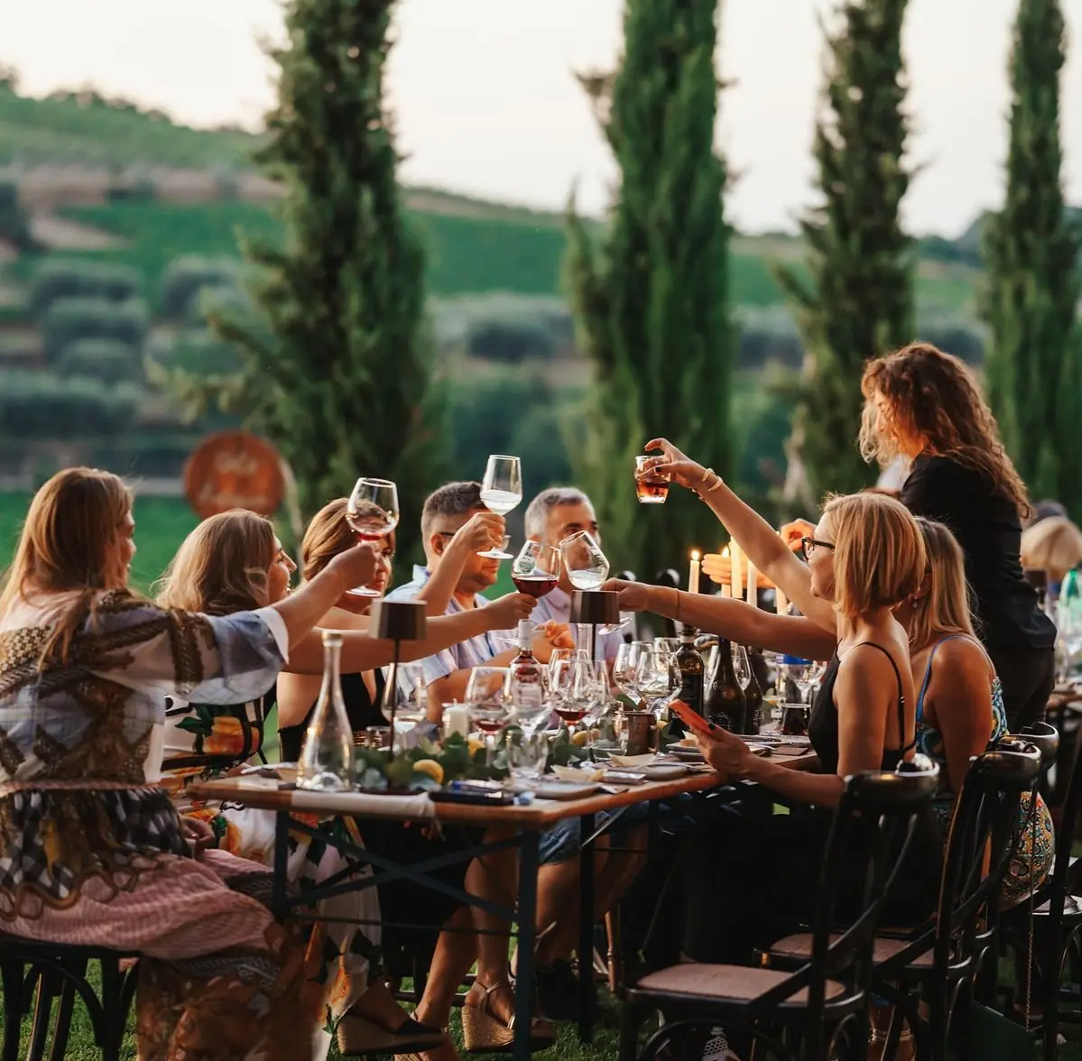 Guests around a long table outdoors raise glasses of red wine together at golden hour, with tall cypress trees and vineyard hills behind them.