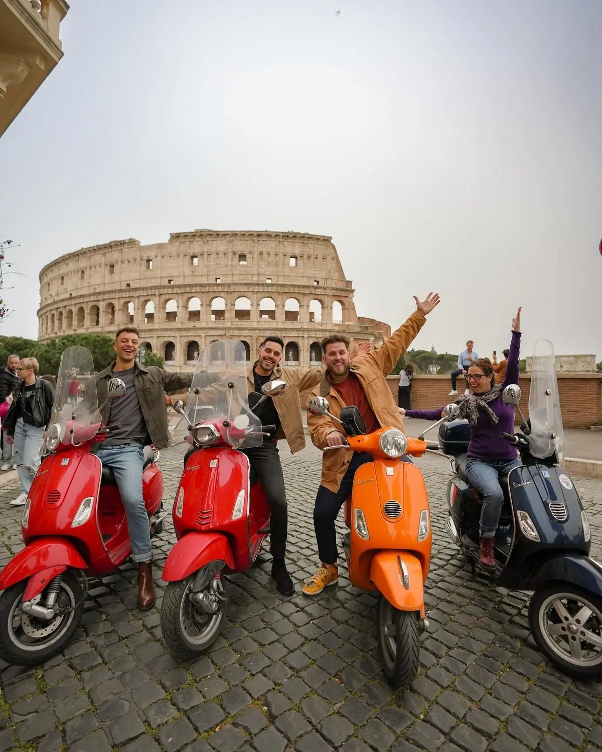 Four smiling travellers sit on red, orange and dark blue Vespa scooters on cobblestones in front of the Colosseum in Rome, two of them with their arms thrown up in the air.