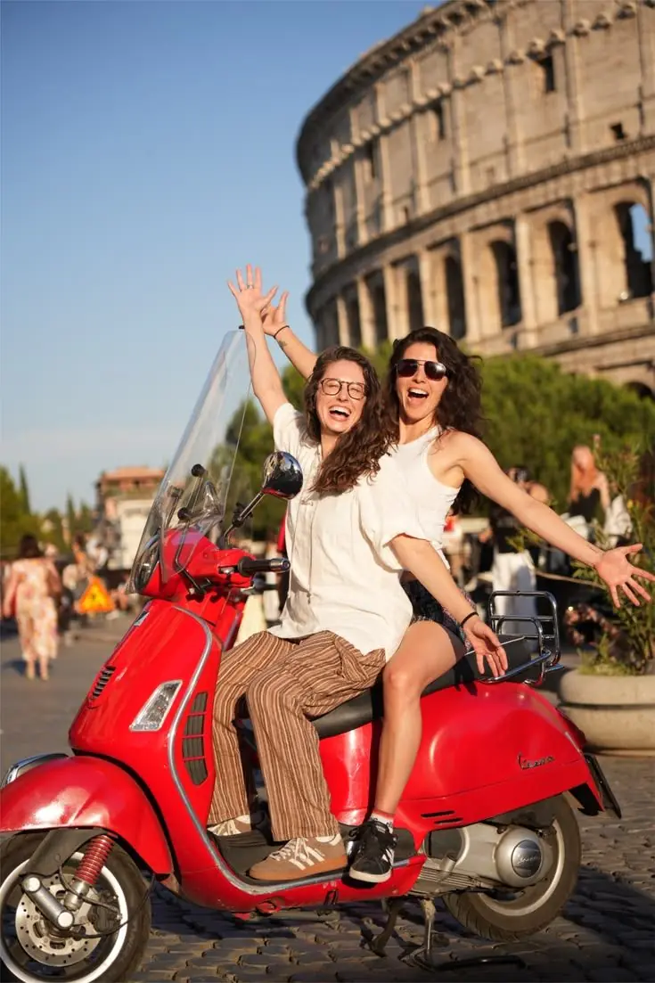 Two women laugh together on a parked red Vespa on a cobbled street, with the Colosseum lit by low sun behind them.